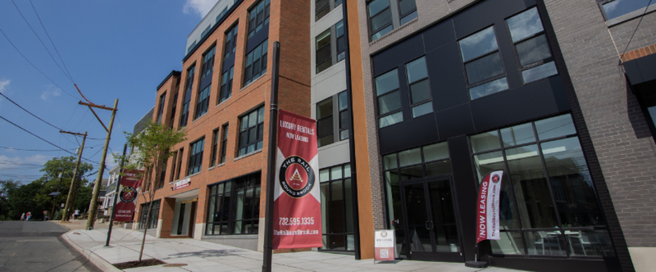 Street-level view of The Rail at Bound Brook luxury apartments featuring leasing banners and modern brick-and-black facade in walkable downtown Bound Brook, New Jersey.